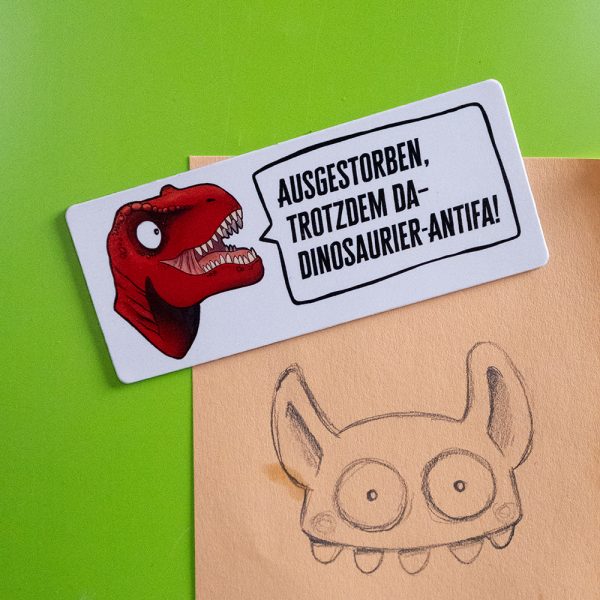 Ein Magnet mit dem Spruch "Ausgestorben, trotzdem da - Dinosaurier-Antifa!" und einem roten Dino-Kopf vor einer grünen Magnetwand
