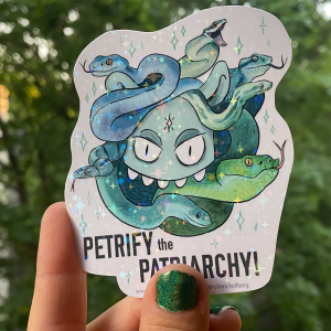 Eine Hand hält einen kontergeschnittenen glitzernden Aufkleber. Darauf ist ein grüner Monsterkopf mit verschiedenen Schlangen als Haaren zu sehen. Darunter steht "Petrify the Patriarchy!"